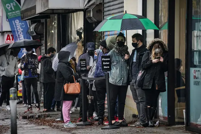 A line of people wearing masks, holding umbrellas, wait on a sidewalk
